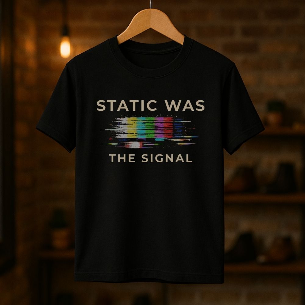 Black t-shirt with 'STATIC WAS THE SIGNAL' text and multicolor static graphic on a wooden hanger against a dark background.
