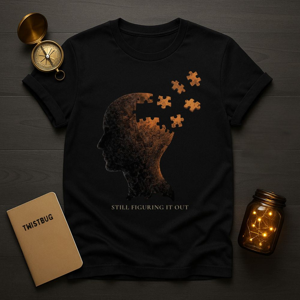 Black graphic T-shirt with a surreal head made of puzzle pieces breaking apart and the quote ‘Still Figuring It Out’, displayed in a dark flat-lay scene with notebook, compass, and cassette
