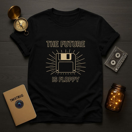 Black “The Future Is Floppy” floppy disk graphic t-shirt laid flat with cassette tape, retro journal, and warm light jar decor.