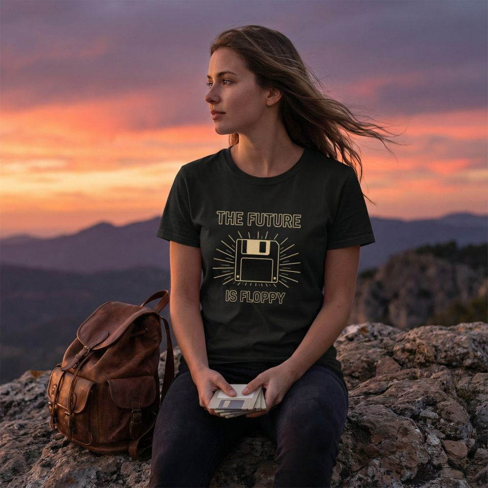 Female model in a black “The Future Is Floppy” floppy disk t-shirt sitting on a rocky cliff at sunset, holding floppy disks with mountains in the background.