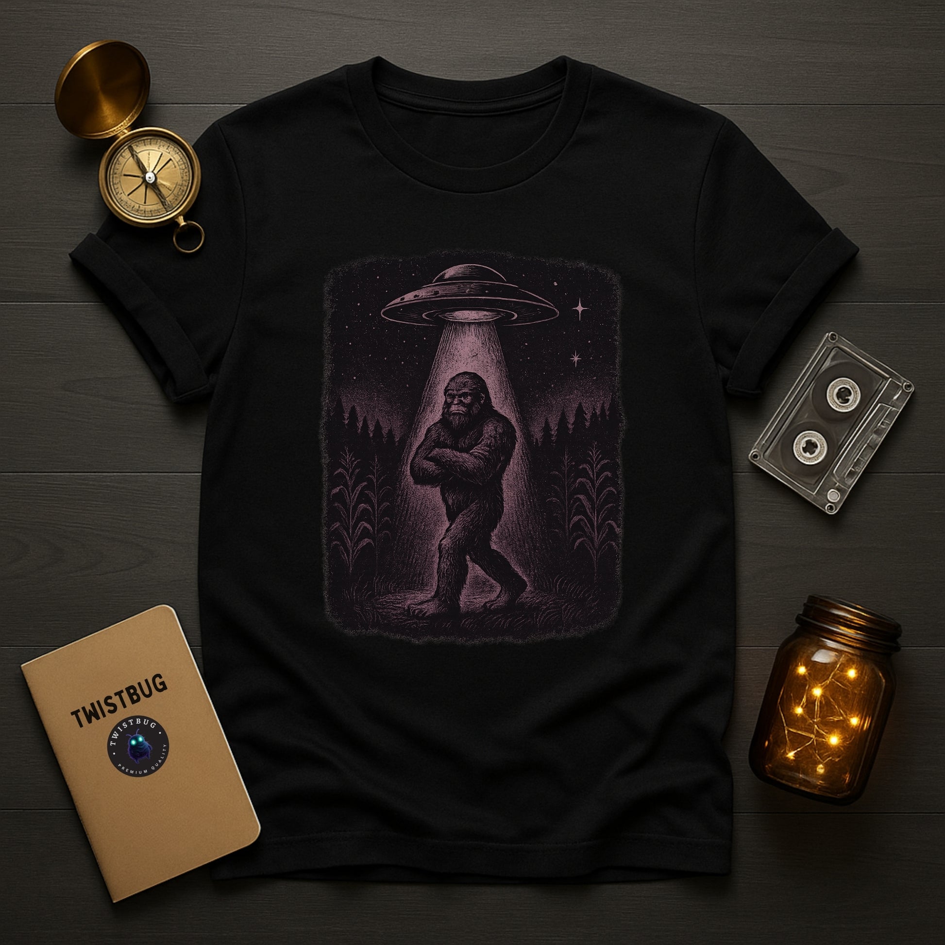 Black t-shirt with a graphic of a Sasquatch under a UFO, placed on a wooden surface with a compass, cassette tape, and jar.