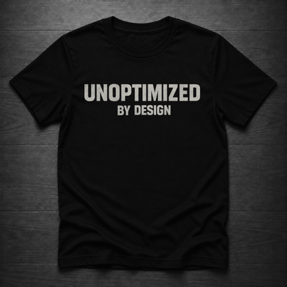 A black crewneck t-shirt with the text 'UNOPTIMIZED BY DESIGN' printed in white across the chest, laid flat on a dark wood plank background.