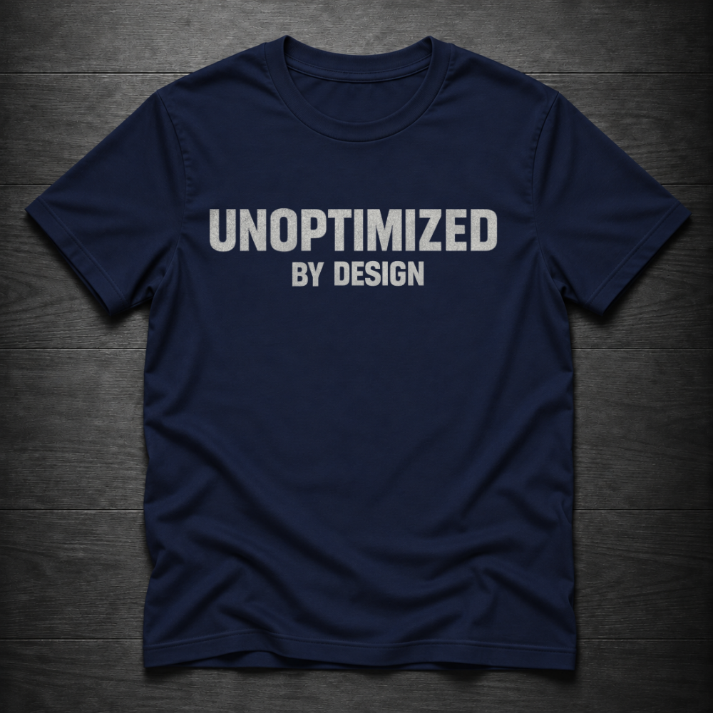 A navy blue short-sleeve t-shirt with the text 'UNOPTIMIZED BY DESIGN' printed in white across the chest, laid flat on a dark wood plank background.