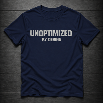 A navy blue short-sleeve t-shirt with the text 'UNOPTIMIZED BY DESIGN' printed in white across the chest, laid flat on a dark wood plank background.