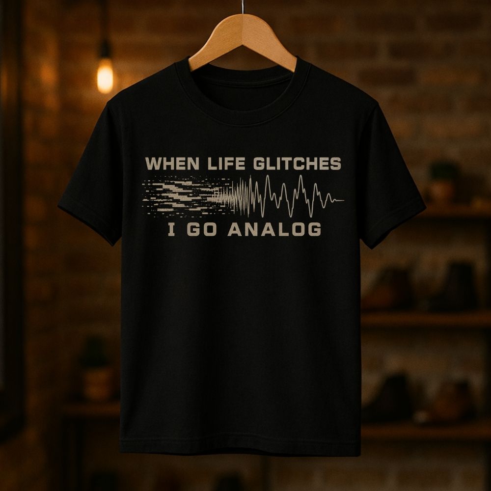 A black short-sleeve graphic t-shirt hanging on a wooden hanger against a warm, blurred brick wall background. The shirt features a distressed beige graphic that reads 'WHEN LIFE GLITCHES' above a pixelated signal wave transitioning into an analog waveform, with 'I GO ANALOG' printed below.