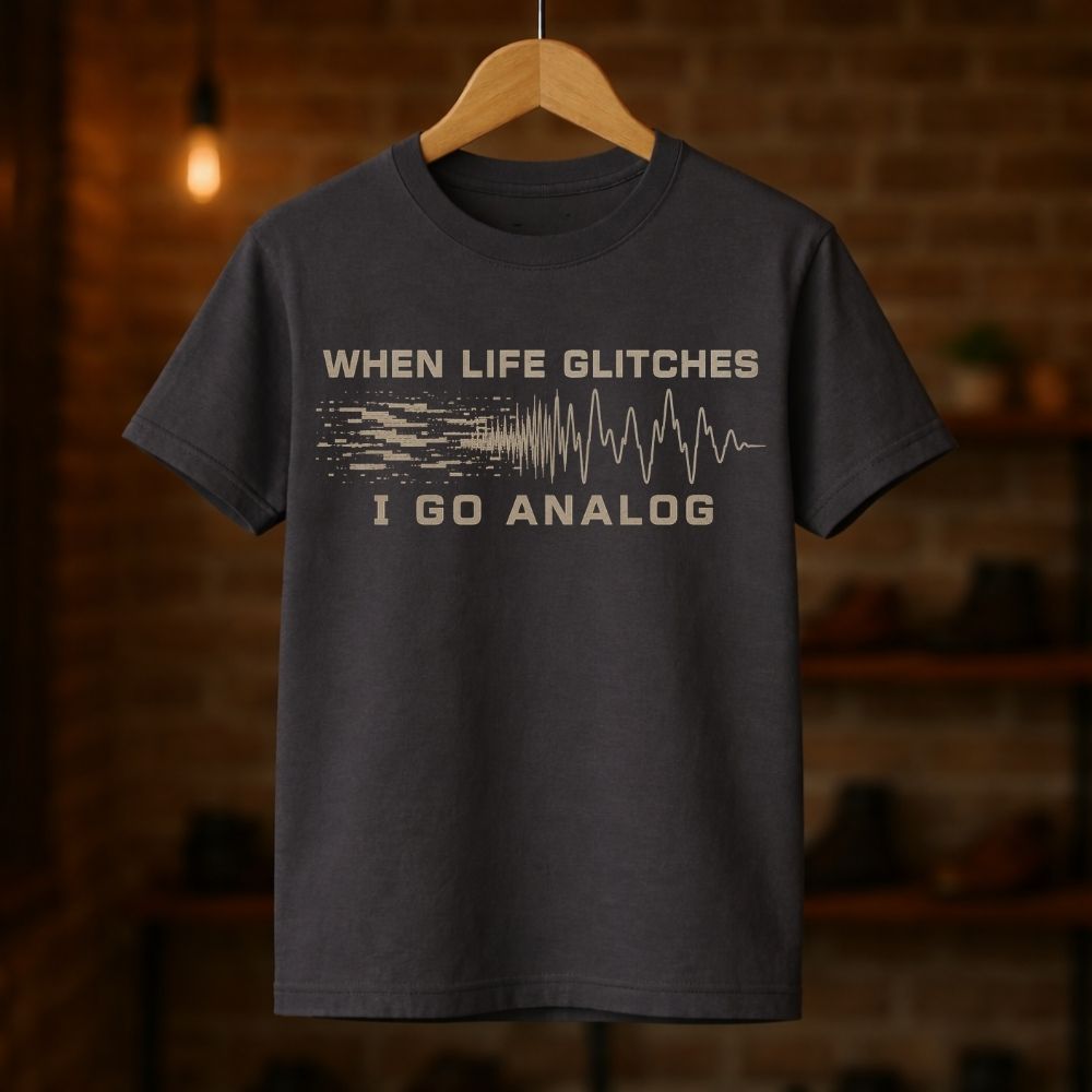 A dark charcoal gray graphic t-shirt hanging on a wooden hanger against a blurred brick wall background. The shirt features a beige graphic print that reads 'WHEN LIFE GLITCHES' above a soundwave design and 'I GO ANALOG' below.
