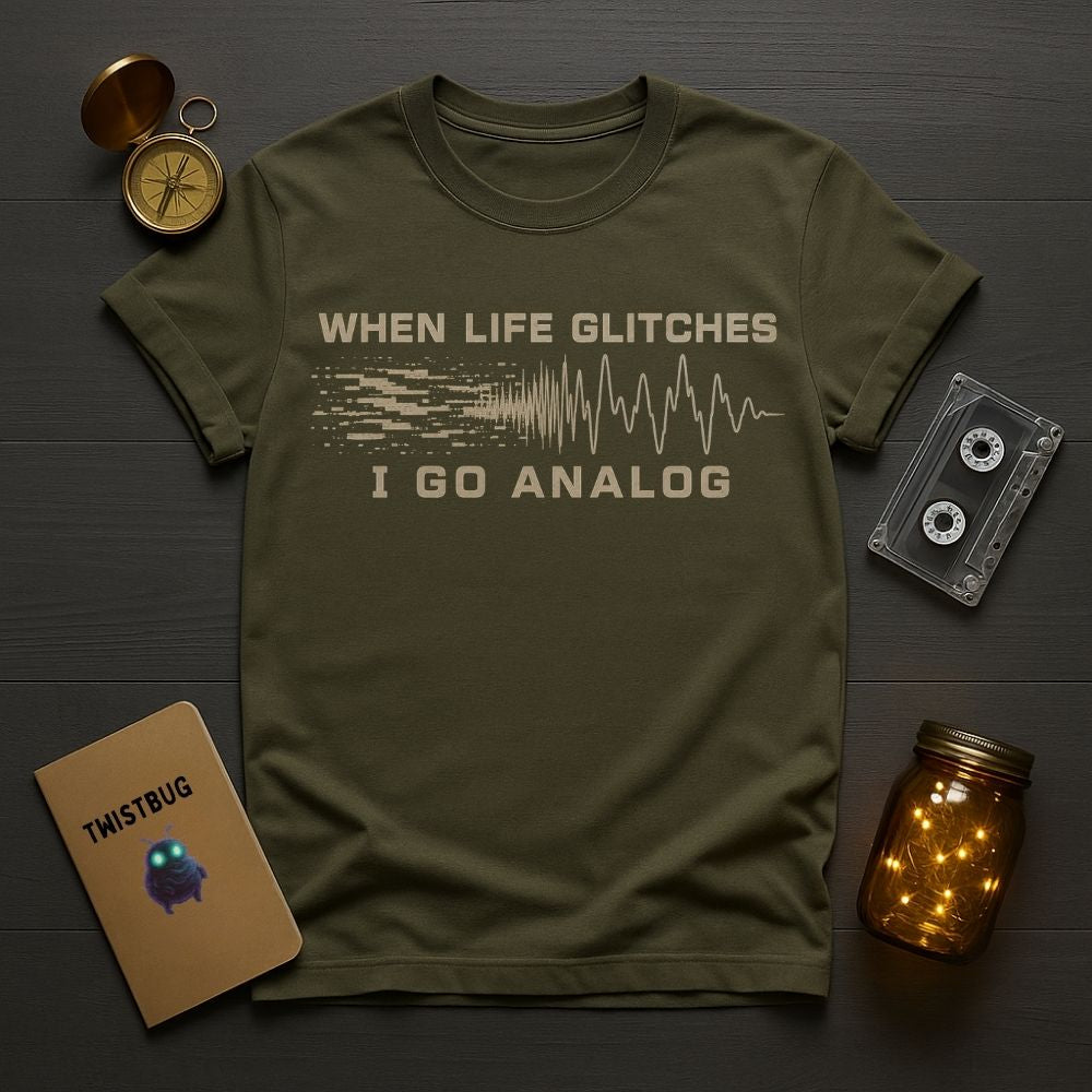 An olive green graphic t-shirt laid flat on a dark wood background, surrounded by cozy props including a vintage compass, a cassette tape, a notebook, and a glowing mason jar. The t-shirt features a beige graphic that reads 'WHEN LIFE GLITCHES I GO ANALOG' with a soundwave design.