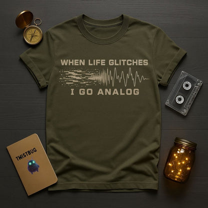 An olive green graphic t-shirt laid flat on a dark wood background, surrounded by cozy props including a vintage compass, a cassette tape, a notebook, and a glowing mason jar. The t-shirt features a beige graphic that reads 'WHEN LIFE GLITCHES I GO ANALOG' with a soundwave design.