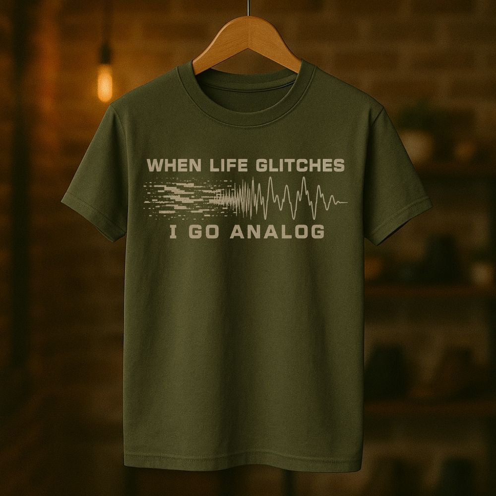 An olive green short-sleeve t-shirt hanging on a wooden hanger against a warm, blurred brick wall background. The shirt features a distressed graphic print in a light beige/cream color that reads 'WHEN LIFE GLITCHES' above a pixelated signal wave and 'I GO ANALOG' below.