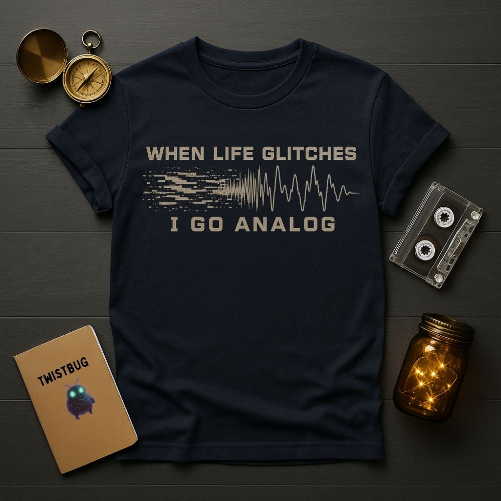 A navy blue graphic t-shirt laid flat on a dark wood background, surrounded by vintage-style props including a compass, cassette tape, notebook, and glowing mason jar. The shirt features a distressed beige graphic that reads 'WHEN LIFE GLITCHES I GO ANALOG' with a pixelated soundwave design.