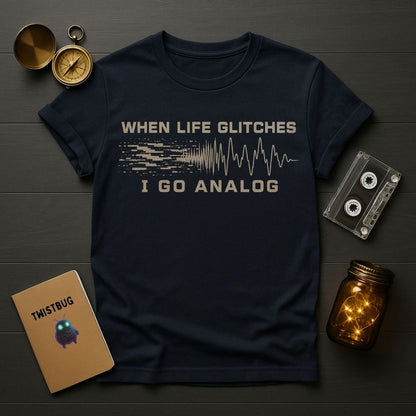A navy blue graphic t-shirt laid flat on a dark wood background, surrounded by vintage-style props including a compass, cassette tape, notebook, and glowing mason jar. The shirt features a distressed beige graphic that reads 'WHEN LIFE GLITCHES I GO ANALOG' with a pixelated soundwave design.