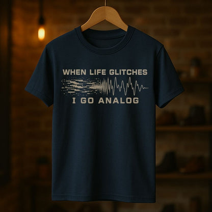 A navy blue short-sleeve t-shirt hanging on a wooden hanger against a warm, blurred background of a brick wall and shelves. The shirt features a graphic print in a light beige/cream color that reads 'WHEN LIFE GLITCHES' above a pixelated signal wave transitioning into an analog waveform, with 'I GO ANALOG' printed below.