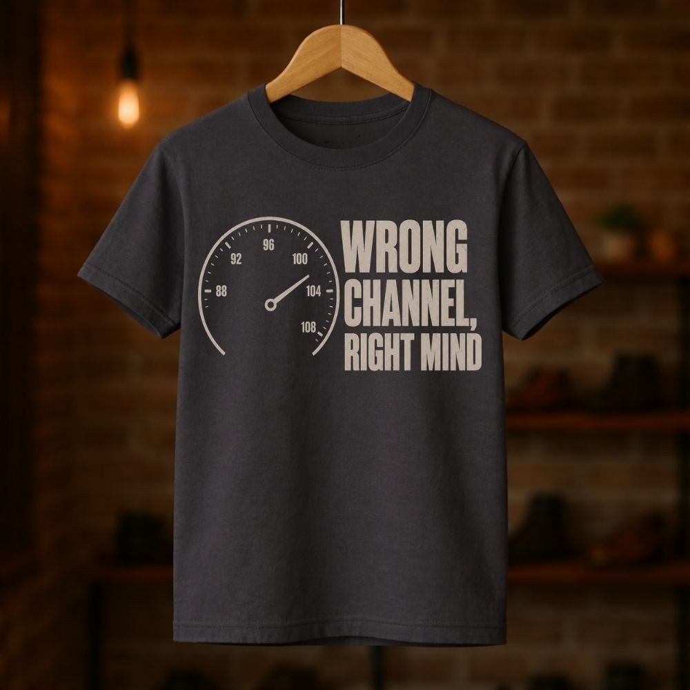A dark charcoal gray t-shirt hanging on a wooden hanger against a blurred brick wall background. The shirt features a graphic print of a speedometer on the left and the text 'WRONG CHANNEL, RIGHT MIND' in a bold, off-white sans-serif font on the right.