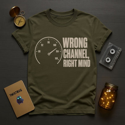 An olive green graphic t-shirt featuring a speedometer and the text 'WRONG CHANNEL, RIGHT MIND' in a distressed cream font. The t-shirt is styled in a flat-lay arrangement on a dark wood background, surrounded by a vintage compass, a notebook labeled 'TWISTBUG', a cassette tape, and a glowing mason jar.