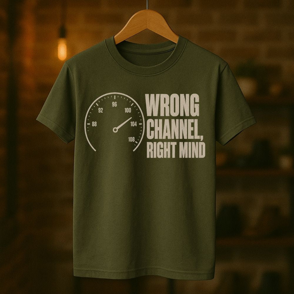 An olive green t-shirt hanging on a wooden hanger against a warm, blurred brick wall background. The shirt features a white graphic print of a speedometer on the left and the text 'WRONG CHANNEL, RIGHT MIND' on the right.
