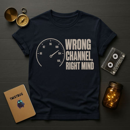 A navy blue graphic t-shirt laid flat on a dark wood background, surrounded by vintage-style props including a pocket watch, cassette tape, notebook, and glowing mason jar. The shirt features a white speedometer graphic and the text 'WRONG CHANNEL, RIGHT MIND'.